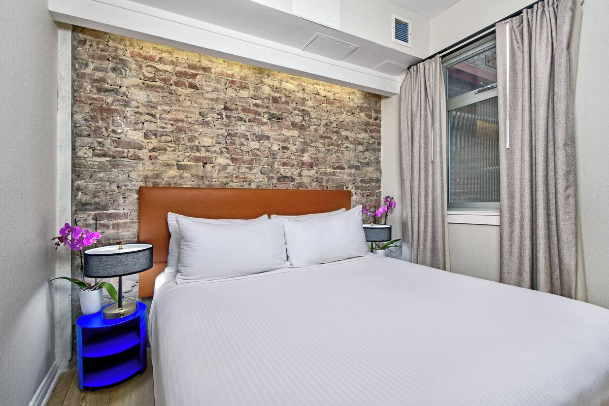 A hotel room with a brick wall, a wooden headboard, a large bed, two bedside tables with lamps, and a window with curtains.