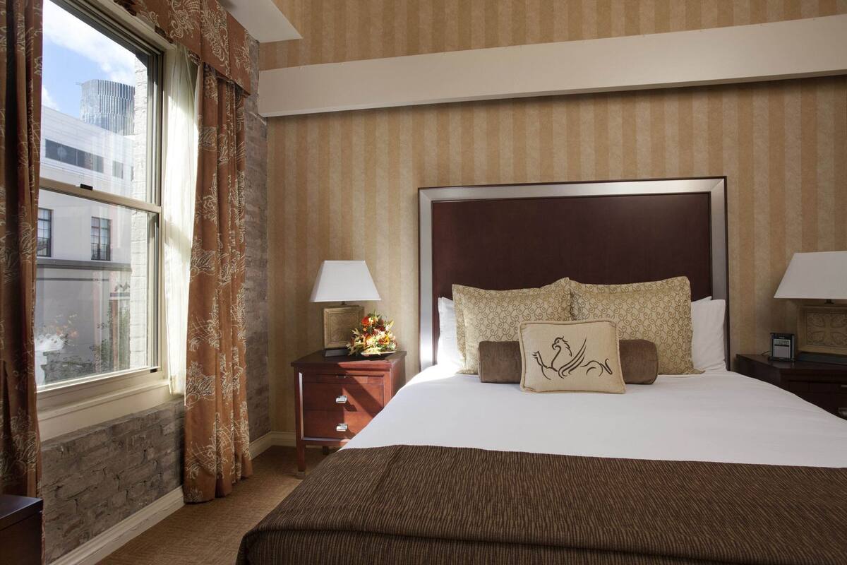 A hotel room with a large bed, a nightstand with a lamp, a window with curtains, and a view of buildings outside.