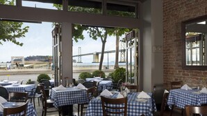 An outdoor dining area with checkered tablecloths, wooden chairs, and a view of a bridge and water.