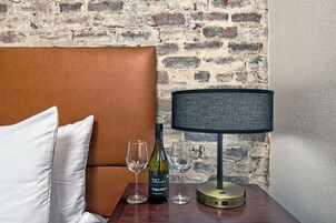 Una habitación de hotel con pared de ladrillo, cabecero marrón, mesita de noche con lámpara, una botella de vino y dos copas.