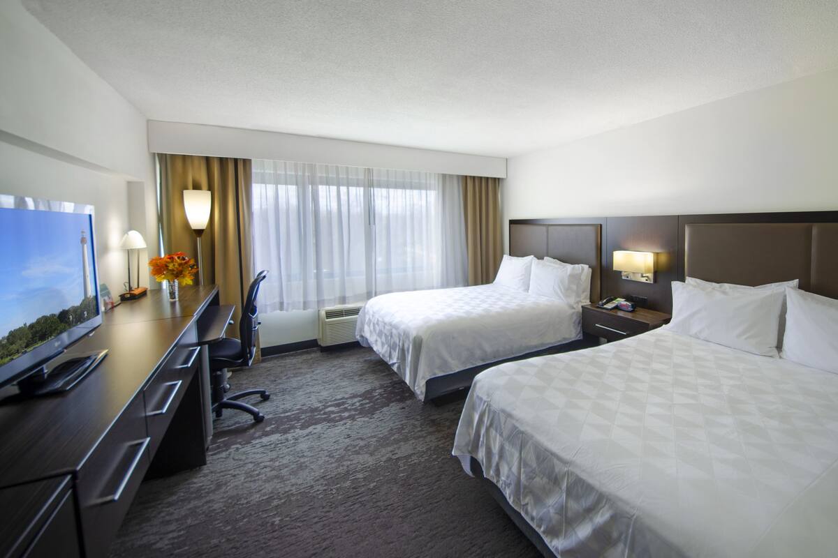 A hotel room with two beds, a desk with a TV, a chair, a lamp, and a window with curtains.