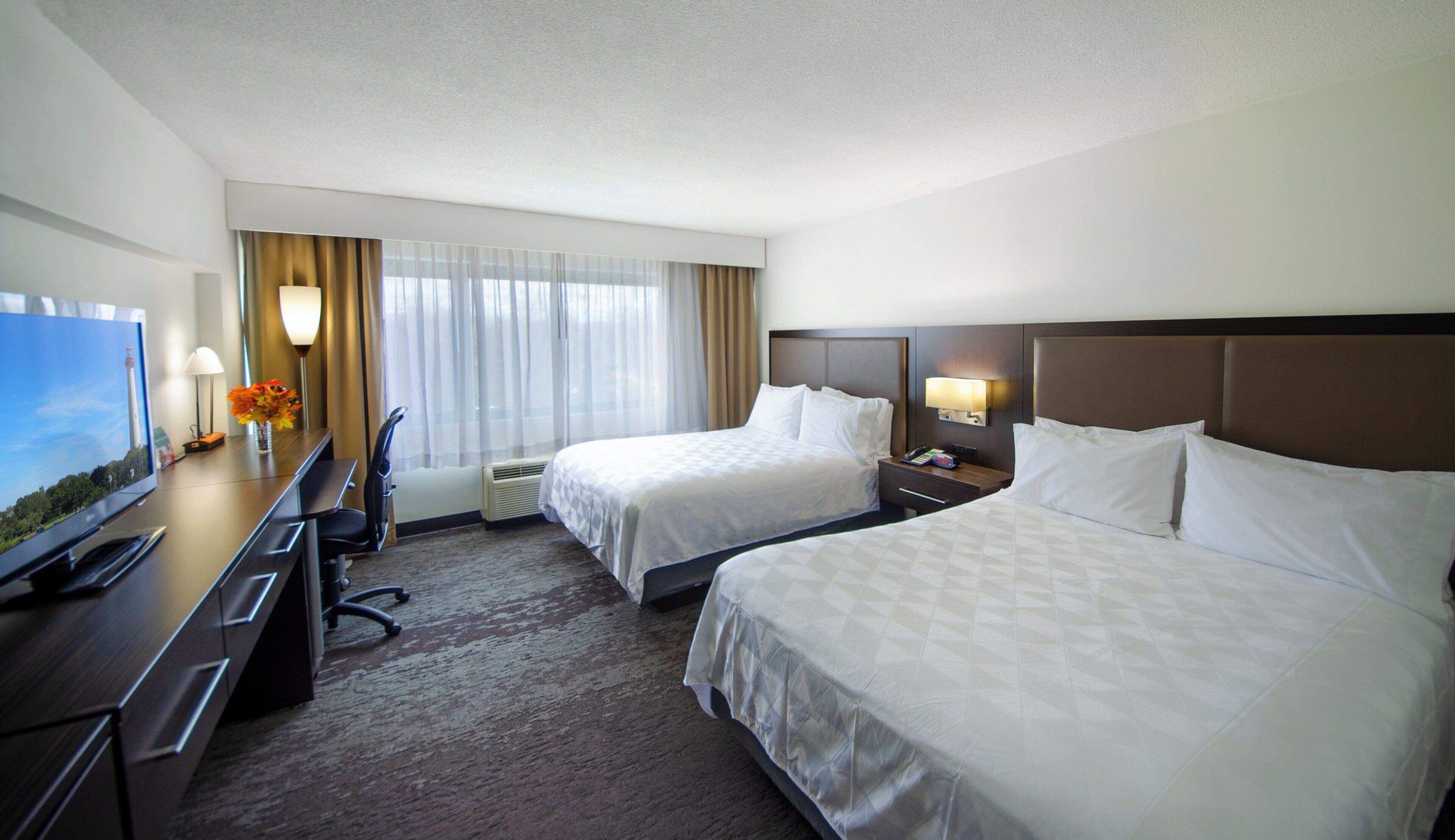 A hotel room with two beds, a desk with a TV, a chair, a desk lamp, and a window with curtains.