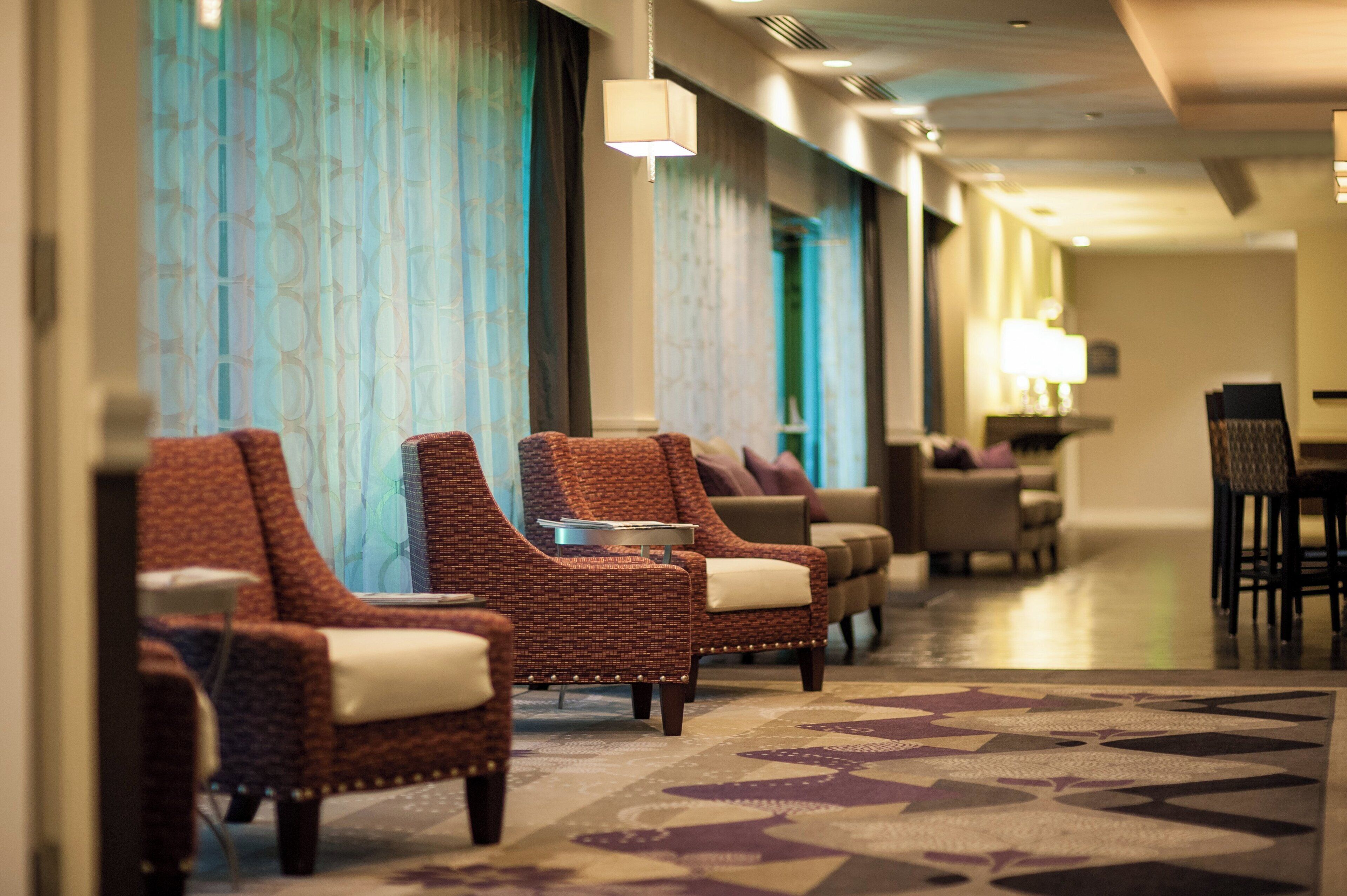 A hotel lobby with a row of wicker armchairs, a patterned carpet, and a dining area in the background.