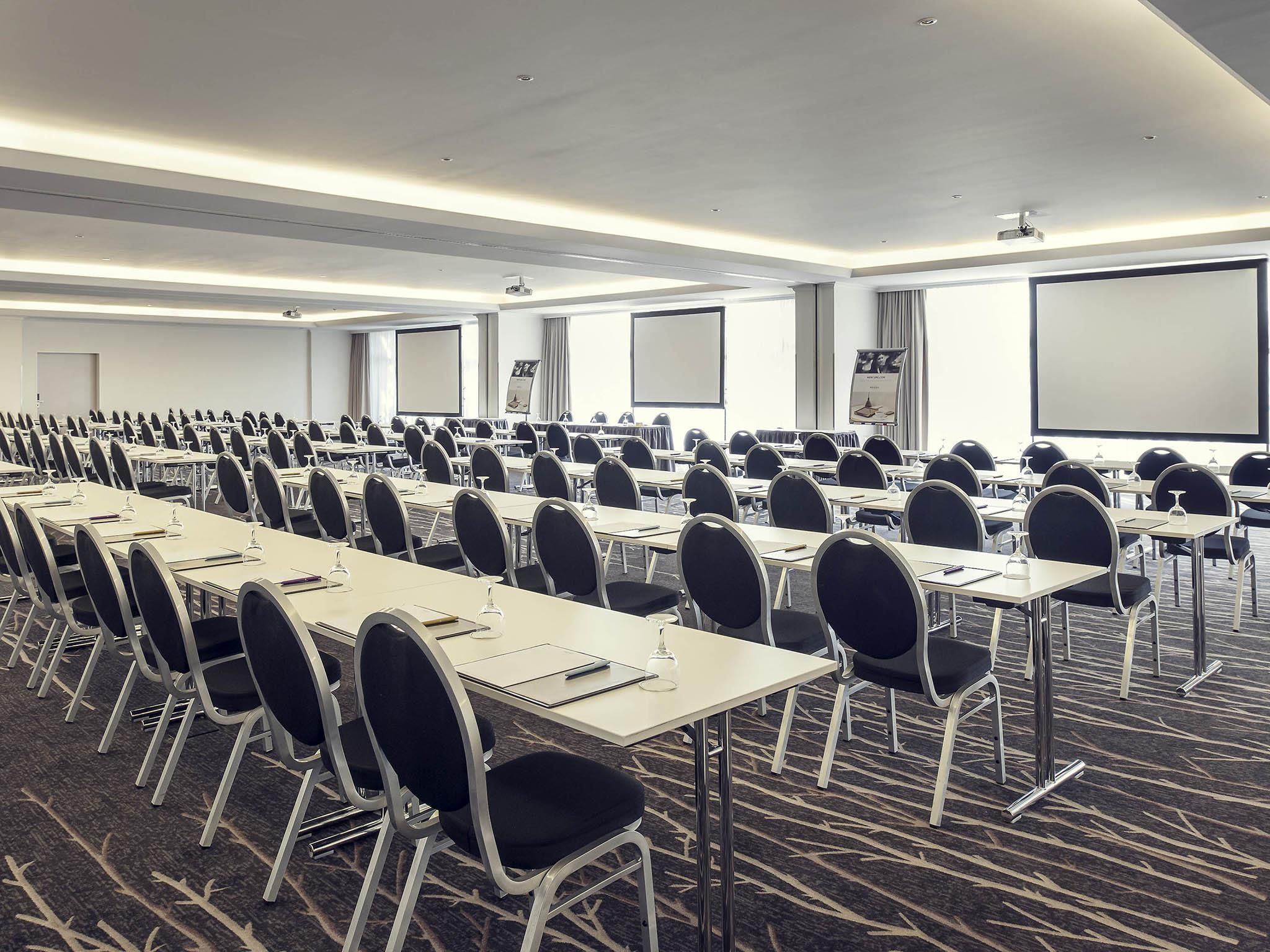 A conference room with rows of tables and chairs, a projector screen, and a carpeted floor.
