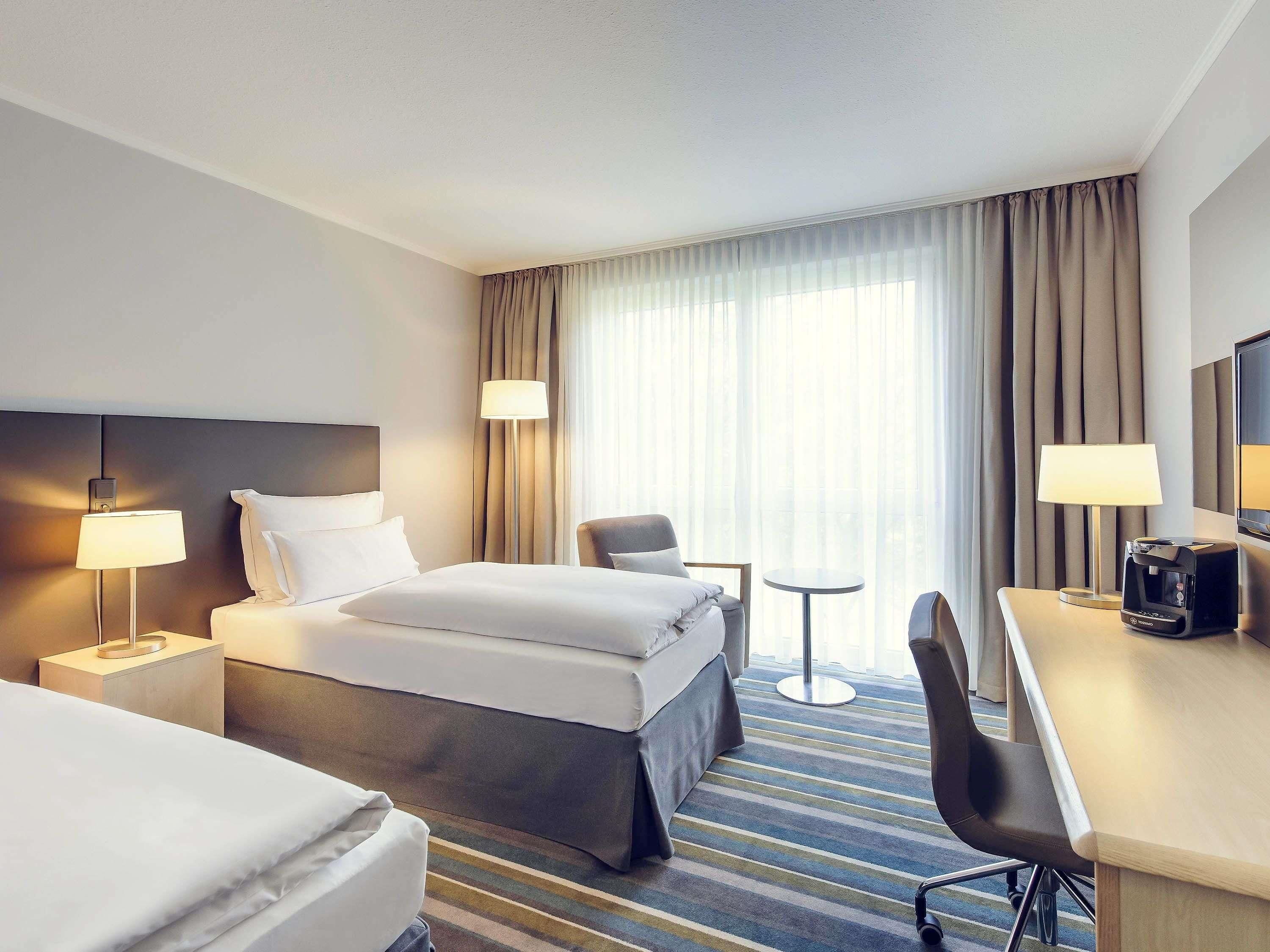A hotel room with a bed, two bedside tables with lamps, a desk with a chair, a coffee maker, and a large window with curtains.