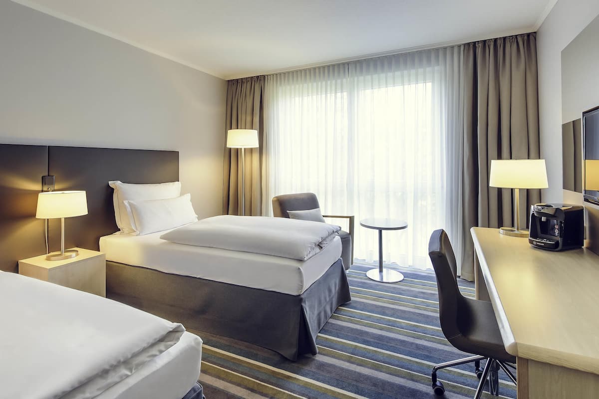 A hotel room with a bed, two bedside tables with lamps, a desk with a chair, a coffee maker, and a view of the outdoors through sheer curtains.