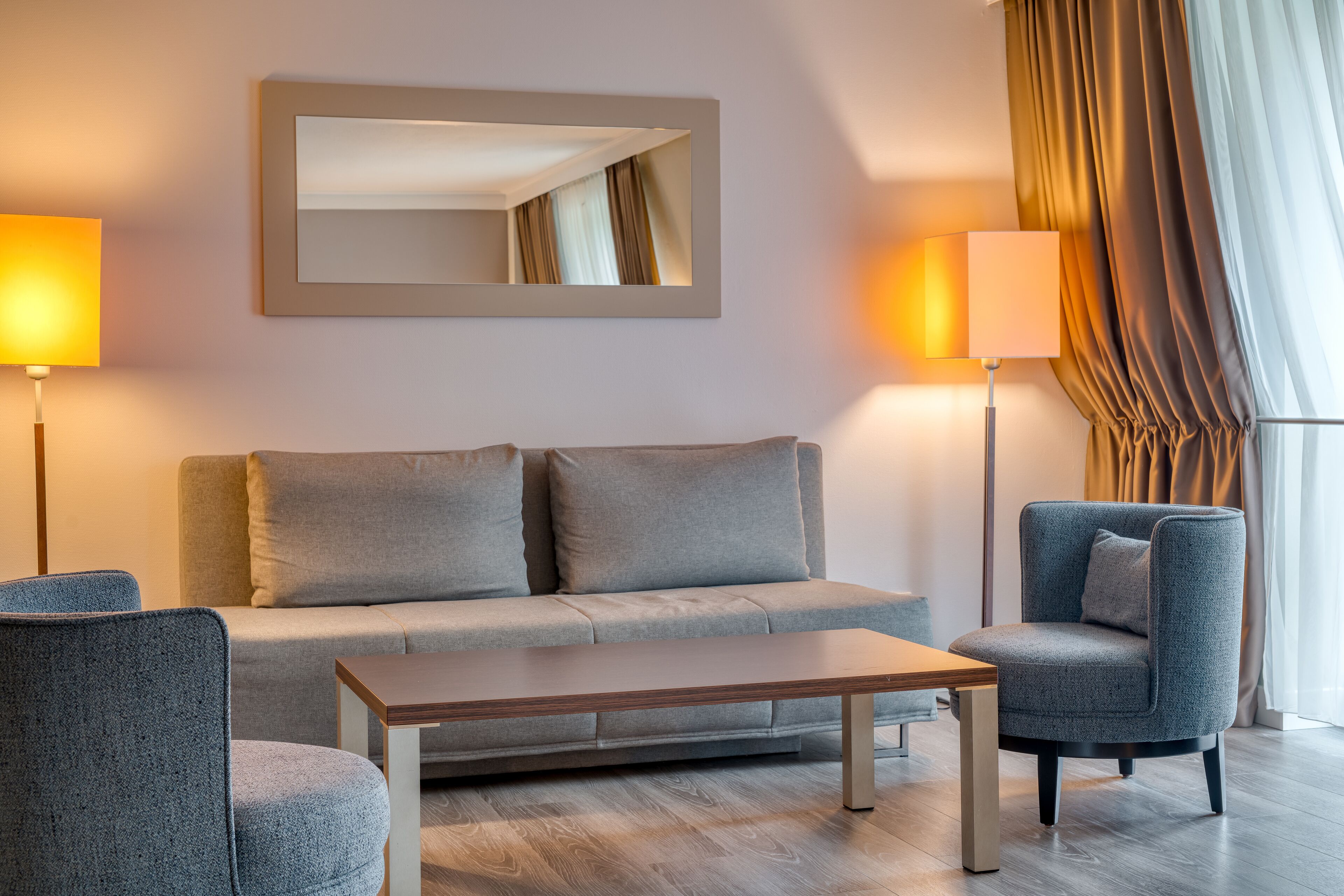 Een moderne woonkamer met een grijze bank, een houten salontafel, een grijze fauteuil en twee vloerlampen.