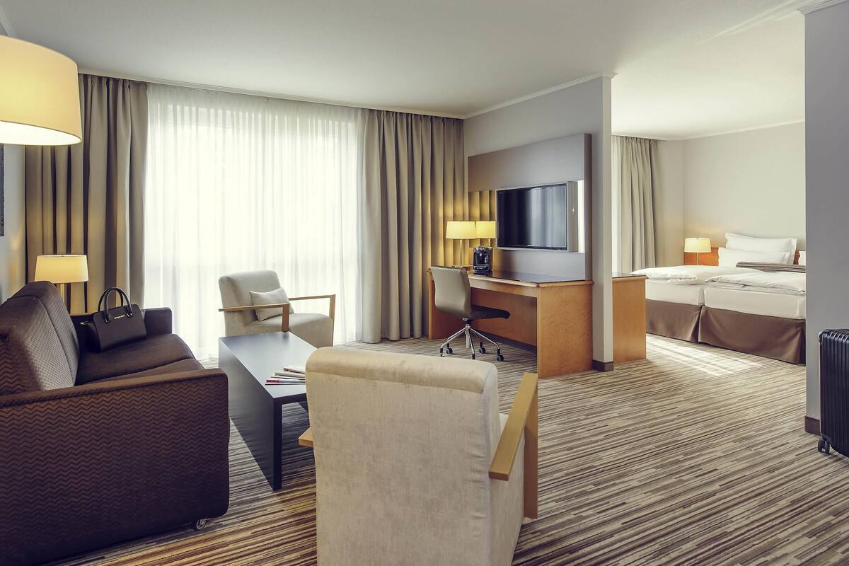 Ein Hotelzimmer mit Sofa, Schreibtisch, Stuhl, Bett, Fernseher und Blick ins Freie.