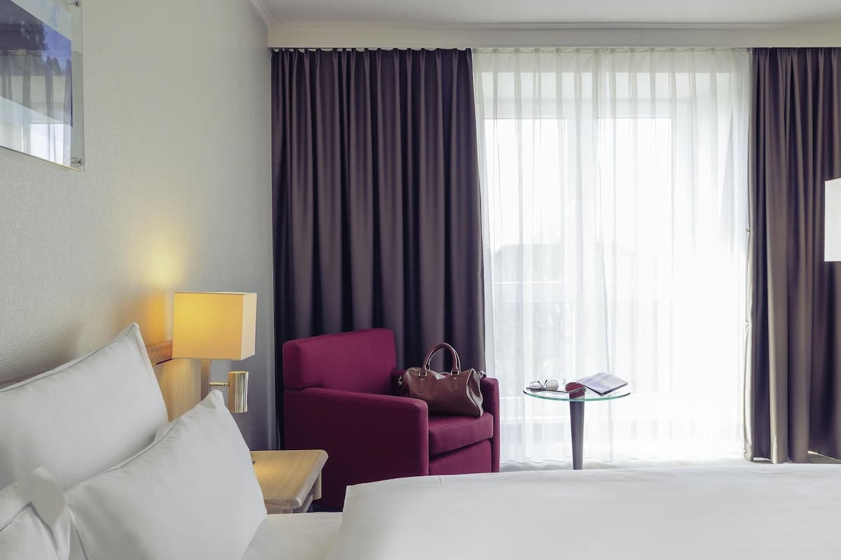 Ein Hotelzimmer mit einem Bett, einem roten Sessel, einem kleinen Tisch mit einer Lampe und Blick auf die Stadt durch große Fenster.