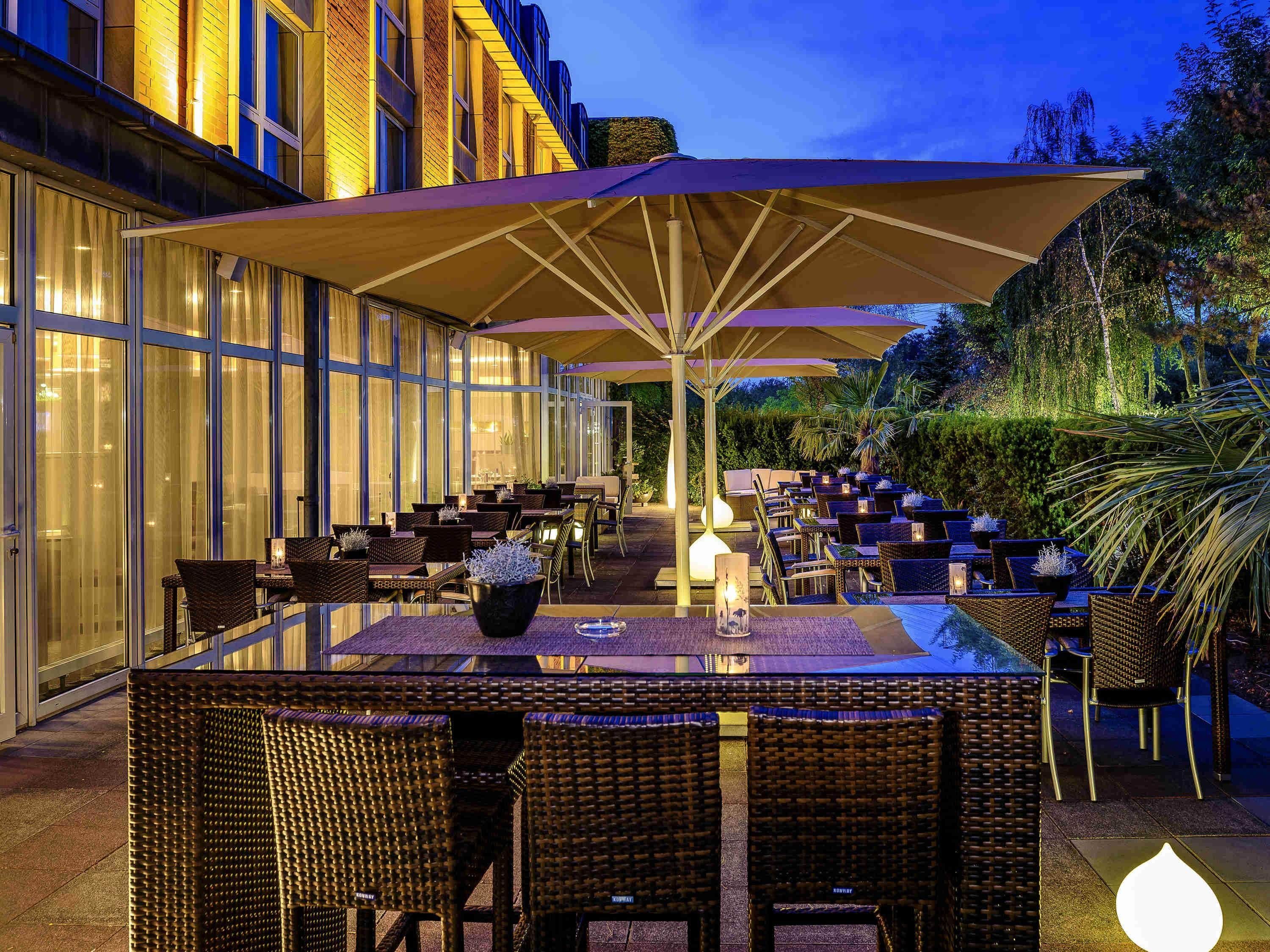 Un espace extérieur aménagé pour les repas, avec des tables et des chaises, de grands parasols, et un bâtiment aux portes vitrées en arrière-plan.