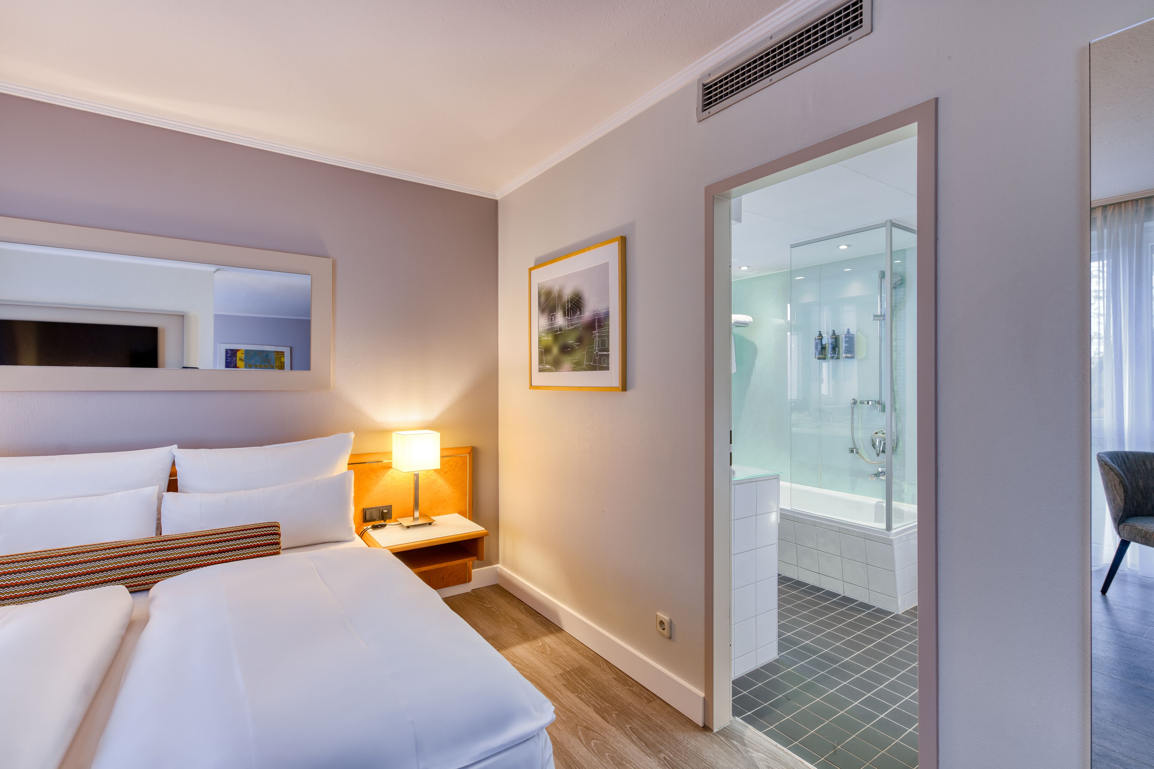 Ein Hotelzimmer mit Bett, Nachttisch, Lampe, gerahmtem Bild und einer Duschkabine.