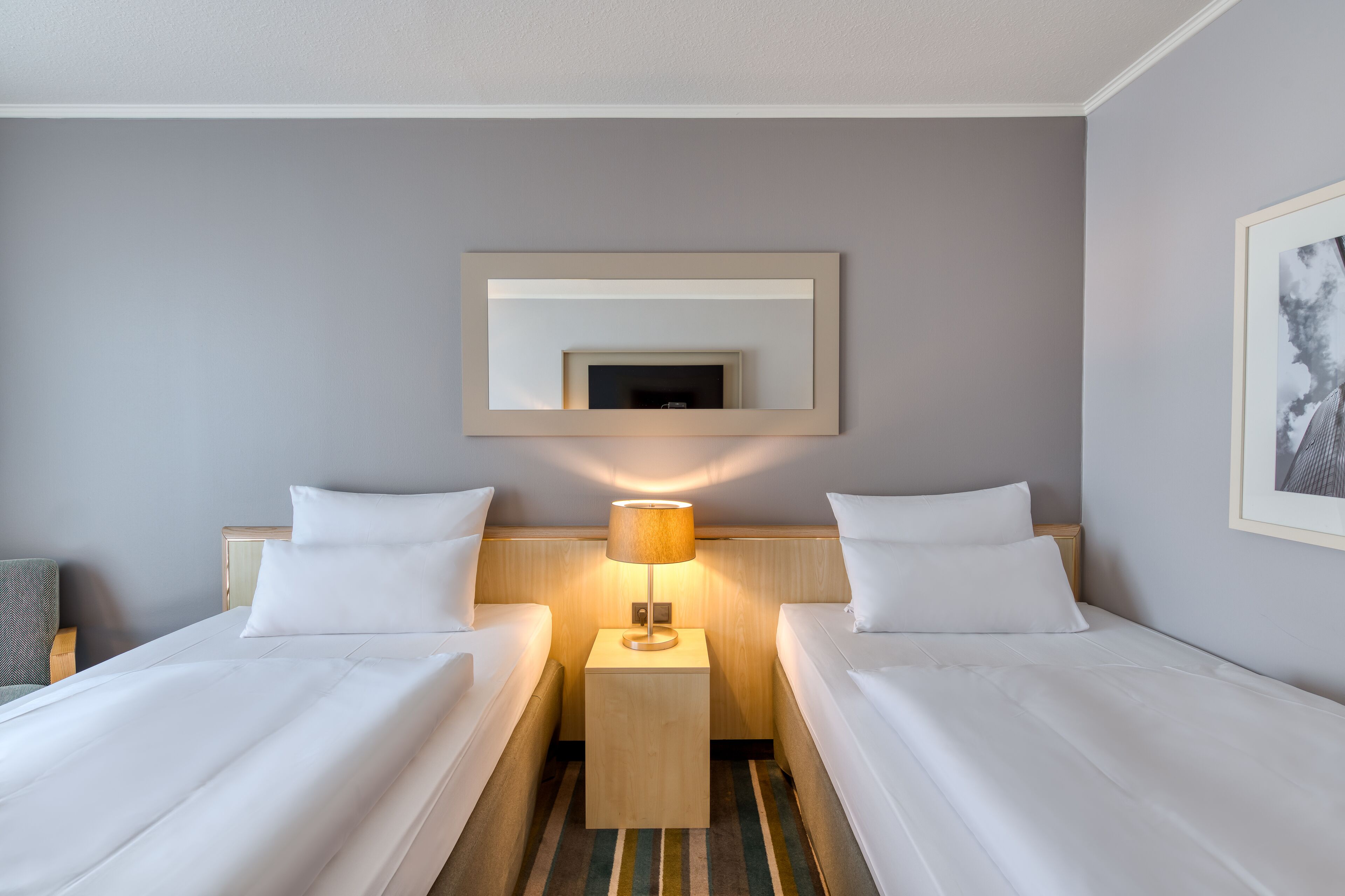 A hotel room with two beds, a bedside table with a lamp, a framed picture on the wall, and a striped rug.