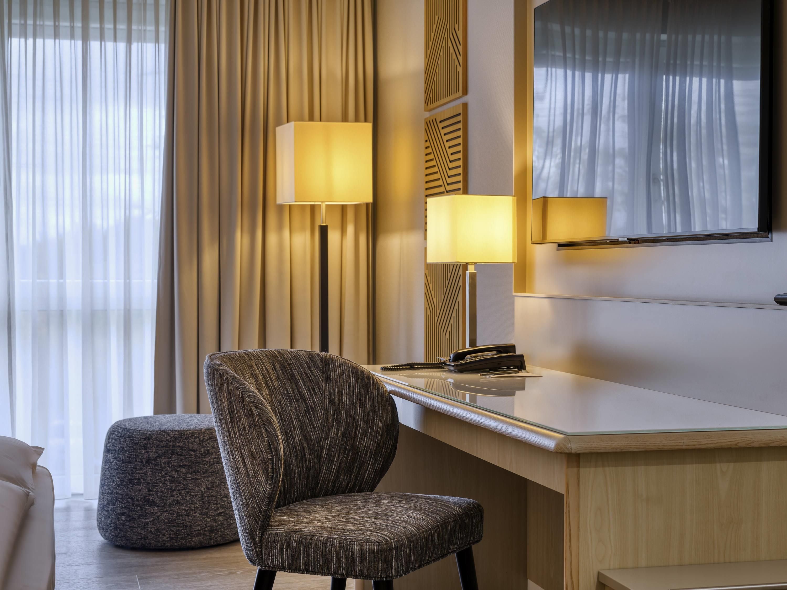 A modern hotel room with a large desk, a comfortable armchair, a round ottoman, and two floor lamps.