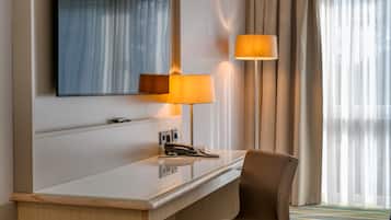 A hotel room with a desk, a mirror, a lamp, a chair, and a window with curtains.