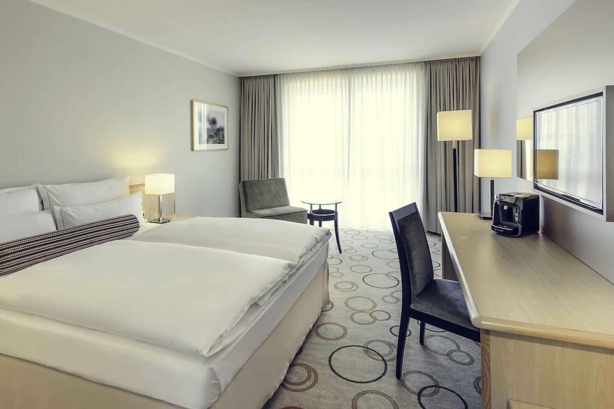 Ein Hotelzimmer mit einem großen Bett, einem Schreibtisch mit Kaffeemaschine, zwei Stühlen, einem kleinen Tisch und einem Fenster mit Vorhängen.