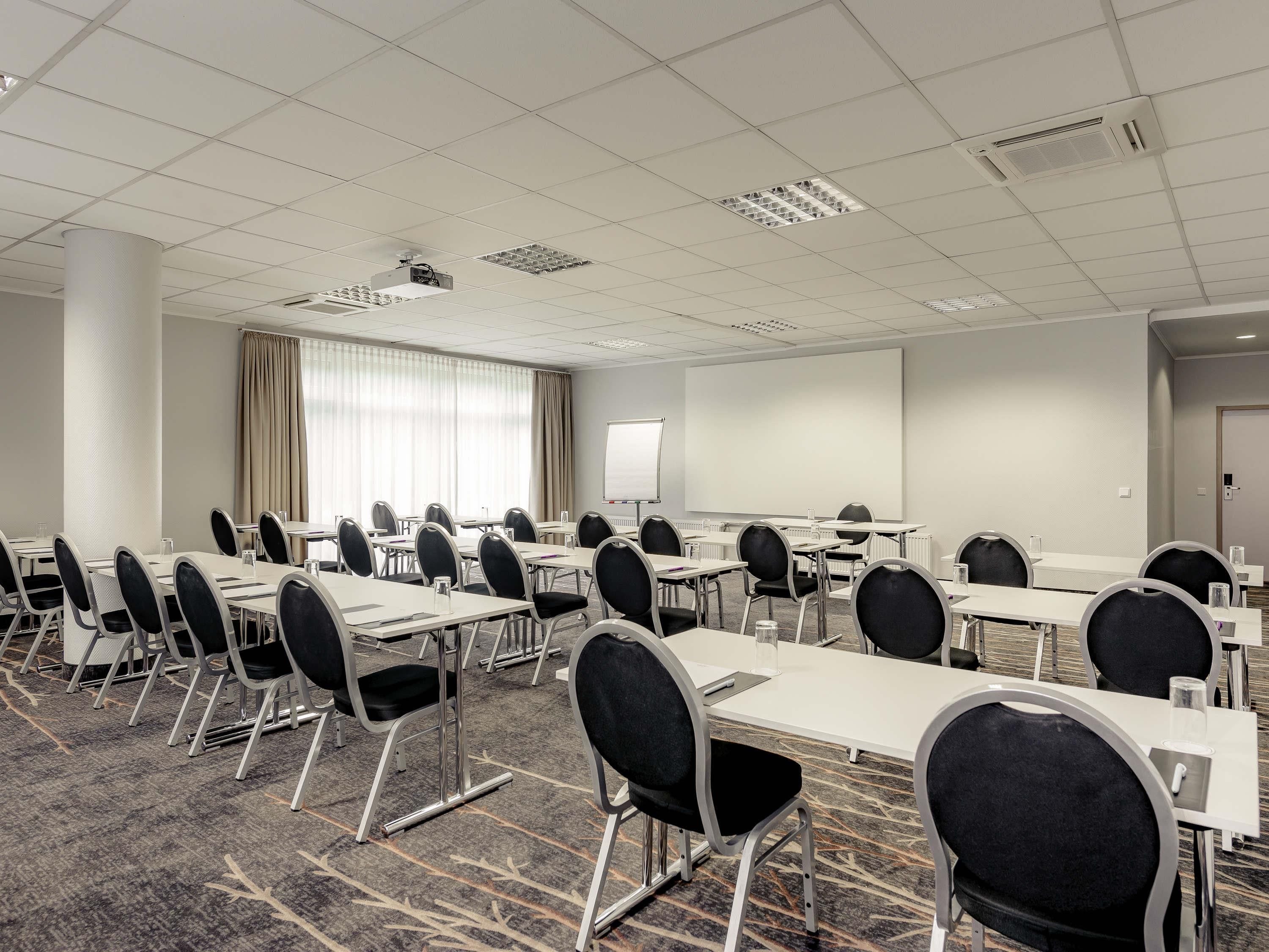 Une salle de conférence équipée de tables et de chaises, d’un projecteur, d’un tableau blanc et d’une grande fenêtre avec des rideaux.