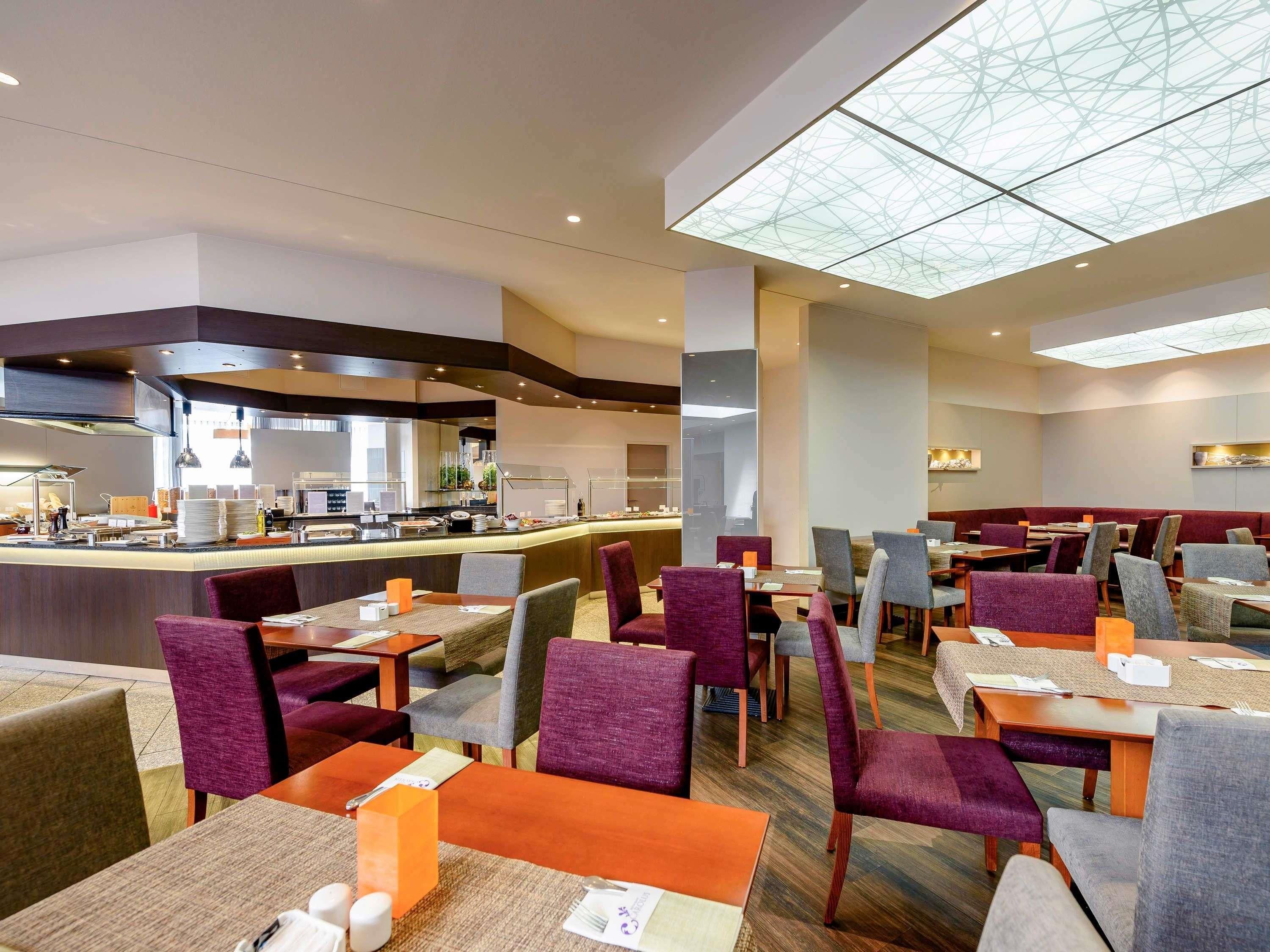 A modern dining area with multiple tables and chairs, a central counter with food display, and a high ceiling with a geometric glass design.
