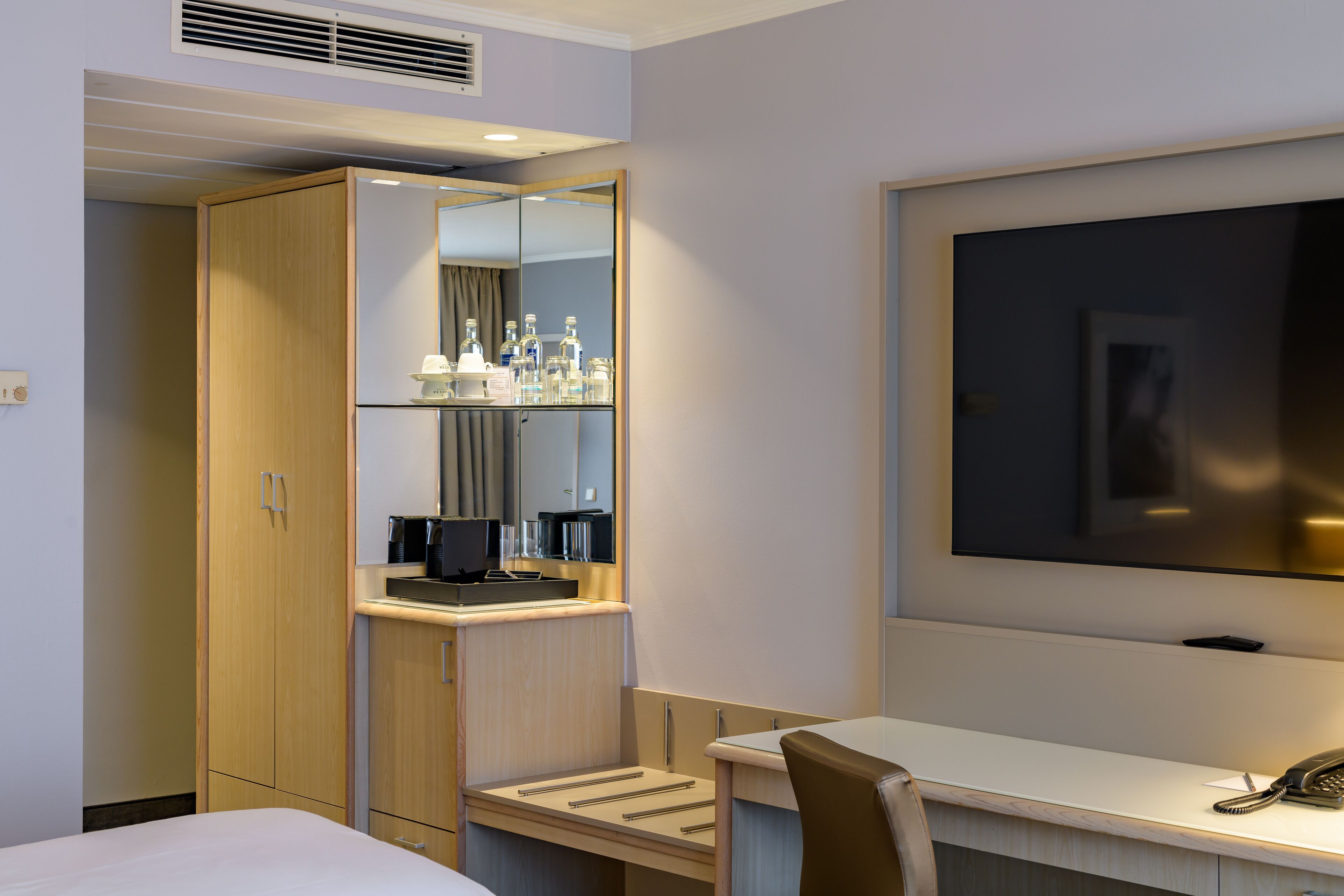 A hotel room with a bed, a desk with a chair, a mirror, a television, and a minibar.