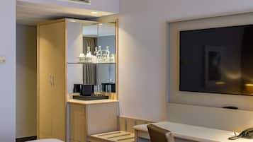 A hotel room with a bed, a desk with a chair, a mirror, a television, and a minibar.