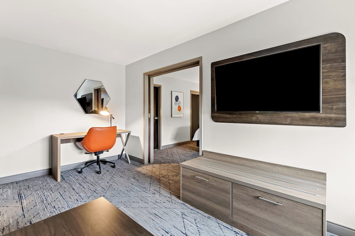 Une chambre d’hôtel moderne avec un bureau en bois, une chaise de bureau orange, un grand miroir et un téléviseur à écran plat fixé au mur.
