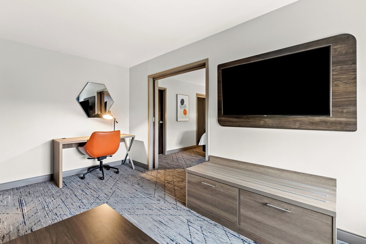 A modern hotel room with a wooden desk, an orange office chair, a large mirror, and a flat-screen TV mounted on the wall.