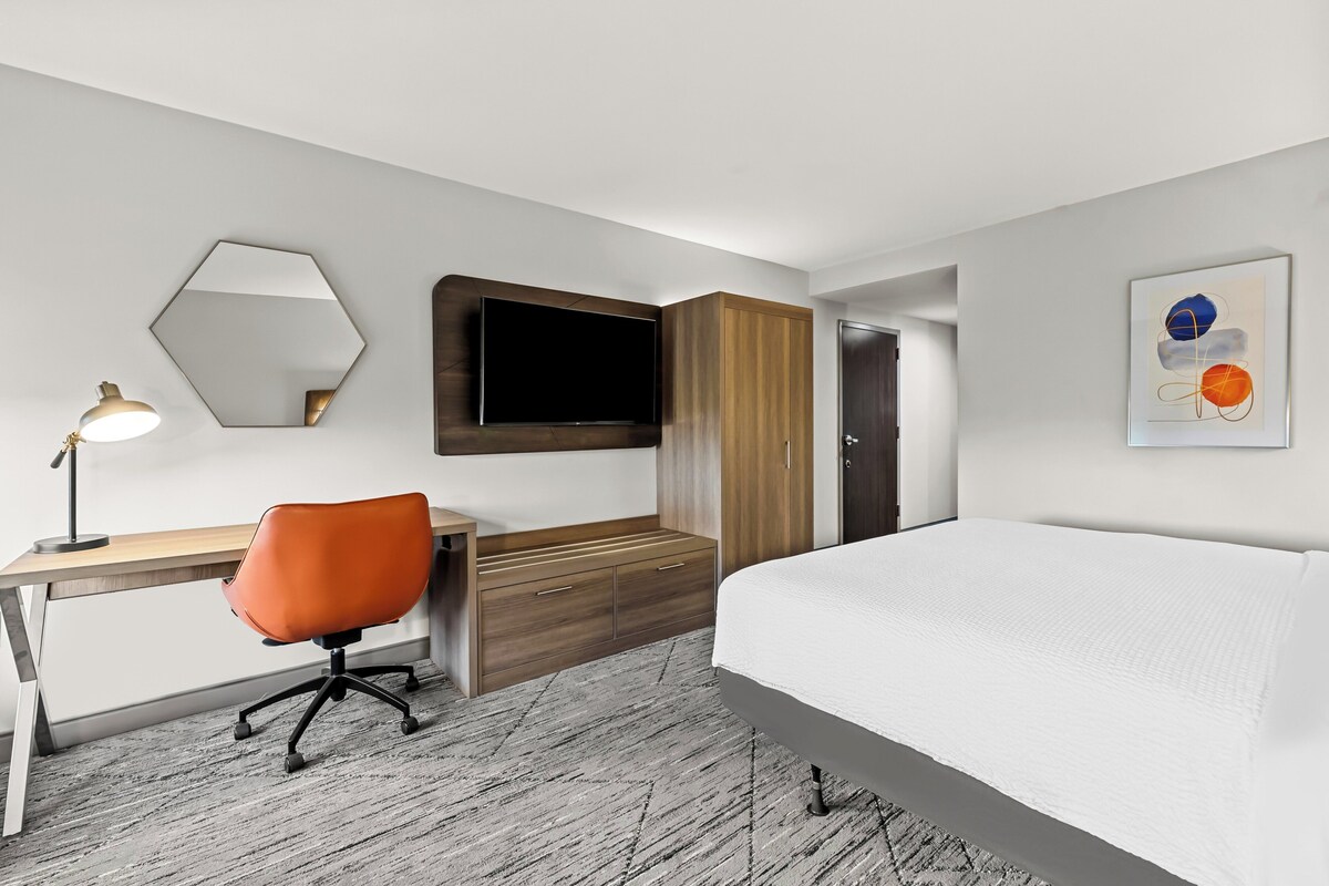 A modern hotel room with a bed, desk, chair, TV, wardrobe, and artwork.