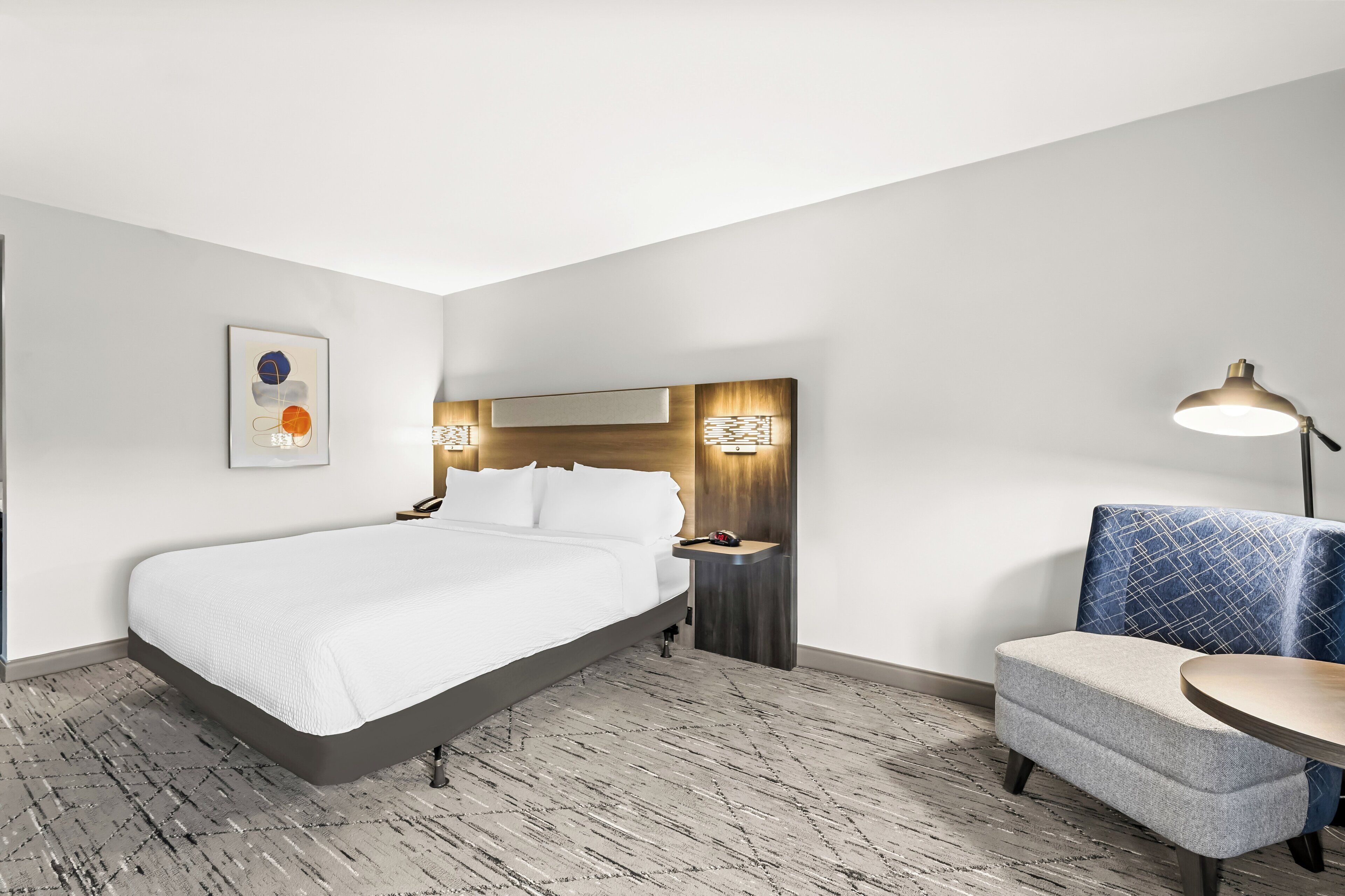 A hotel room with a bed, a chair, a small table, and a wall-mounted lamp.