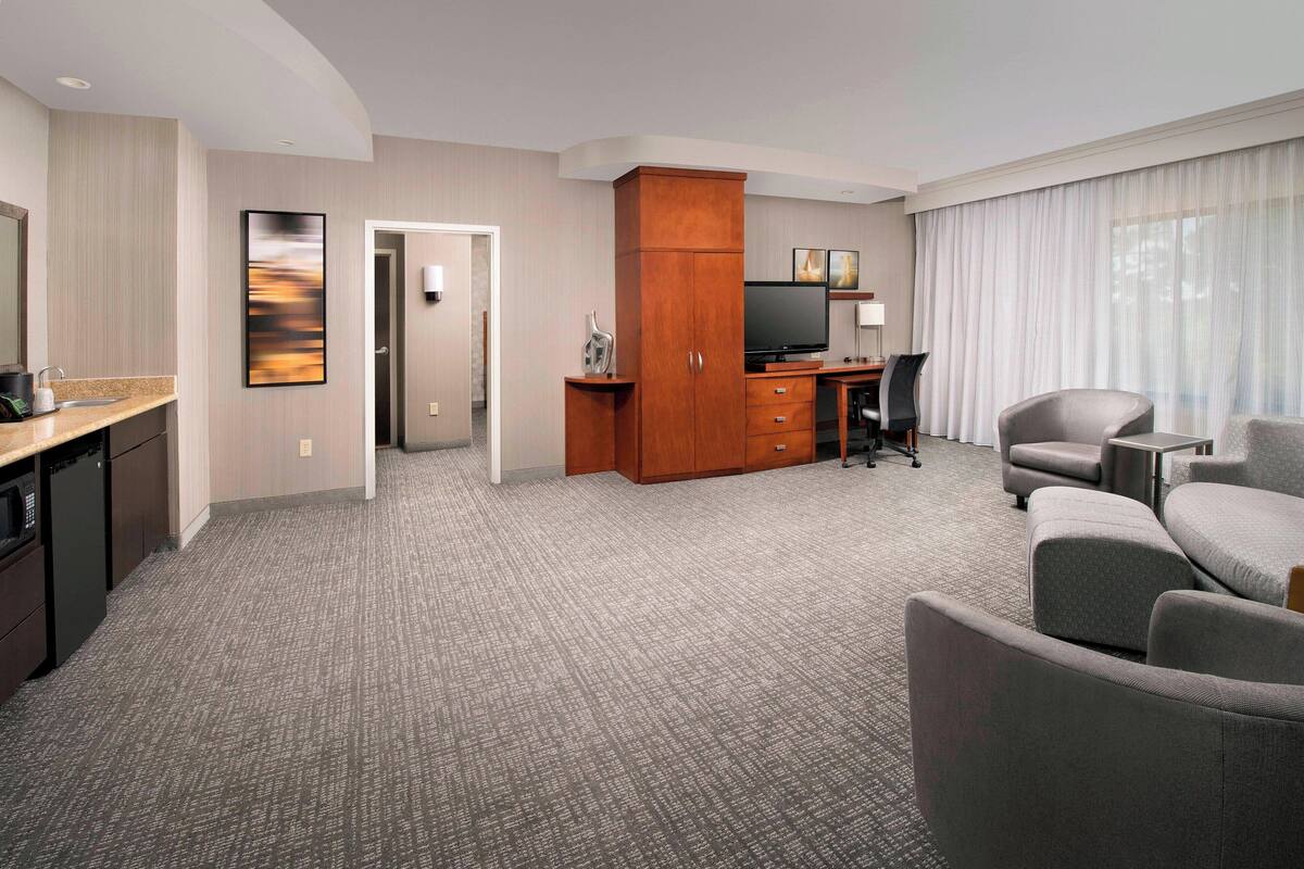 A hotel room with a kitchenette, a flat-screen TV, a desk, and a comfortable seating area with a sofa and armchairs.