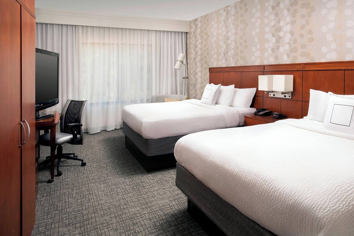 A hotel room with two beds, a desk with a chair, a TV, and a lamp.