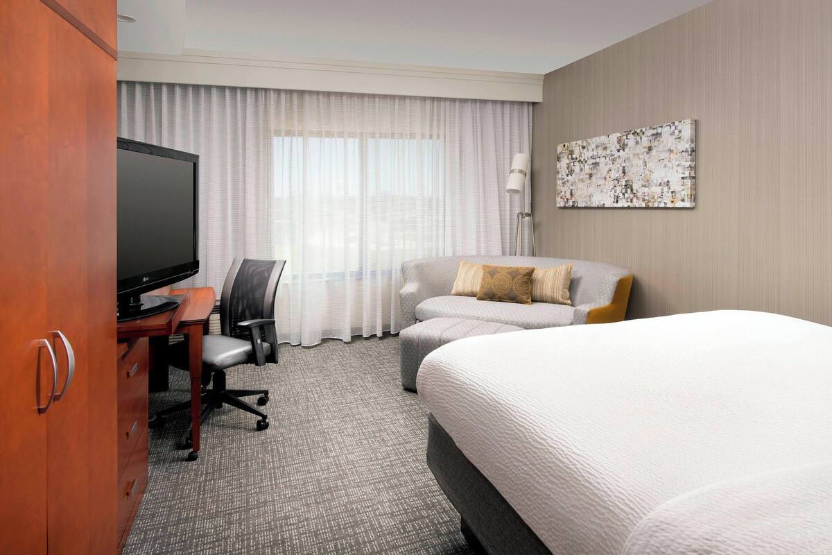 A hotel room with a large bed, a desk with a television, a comfortable armchair, and a large window with curtains.