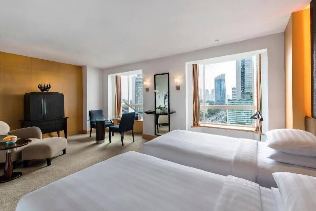 A hotel room with two beds, a desk, a chair, a wardrobe, and a large window with a city view.