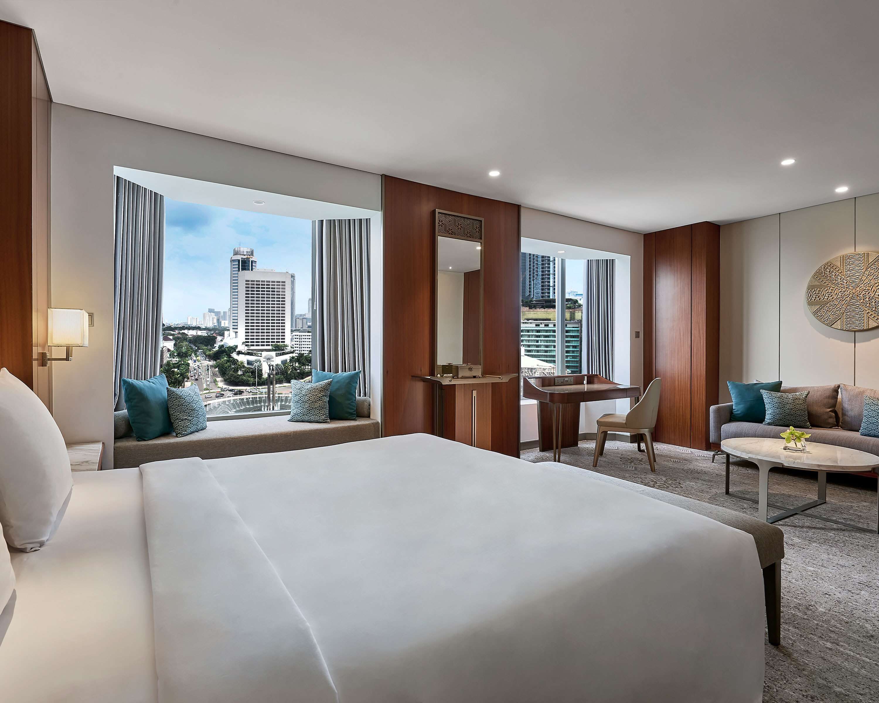 A hotel room with a large bed, a desk, a chair, and a sofa. There are large windows with curtains, a view of the city, and a round wall decoration.