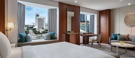 A hotel room with a large bed, a desk, a chair, and a sofa. There are large windows with curtains, a view of the city, and a round wall decoration.