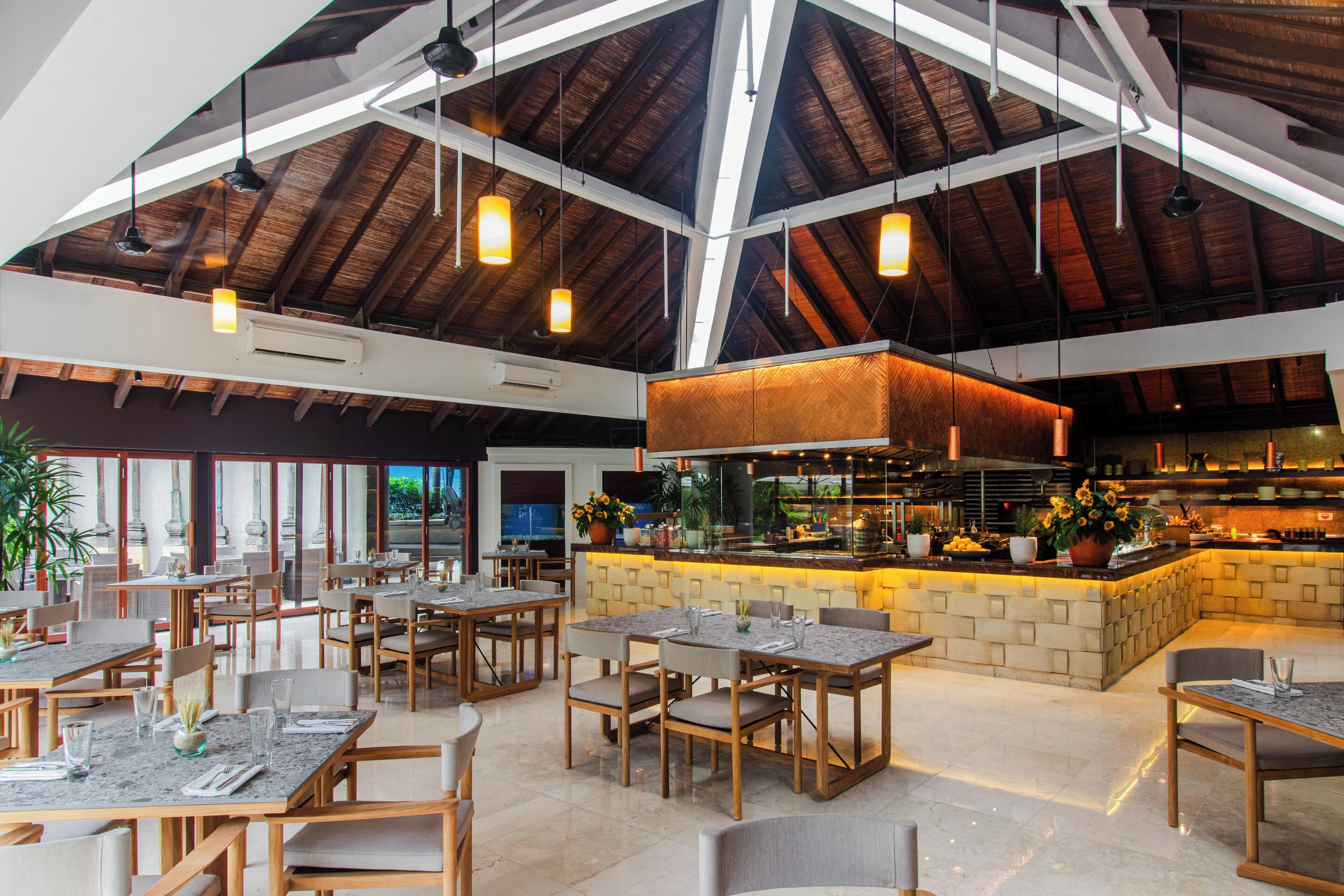 A spacious restaurant with high ceilings, wooden beams, and a central bar area. Tables are arranged for dining, and the space is well-lit with pendant lighting.
