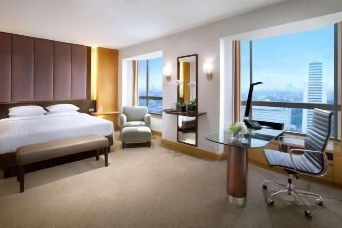 A hotel room with a bed, a desk, a chair, and a large window with a city view.
