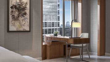 A modern hotel room with a large window, a desk with a lamp, a framed artwork, and a view of the cityscape.