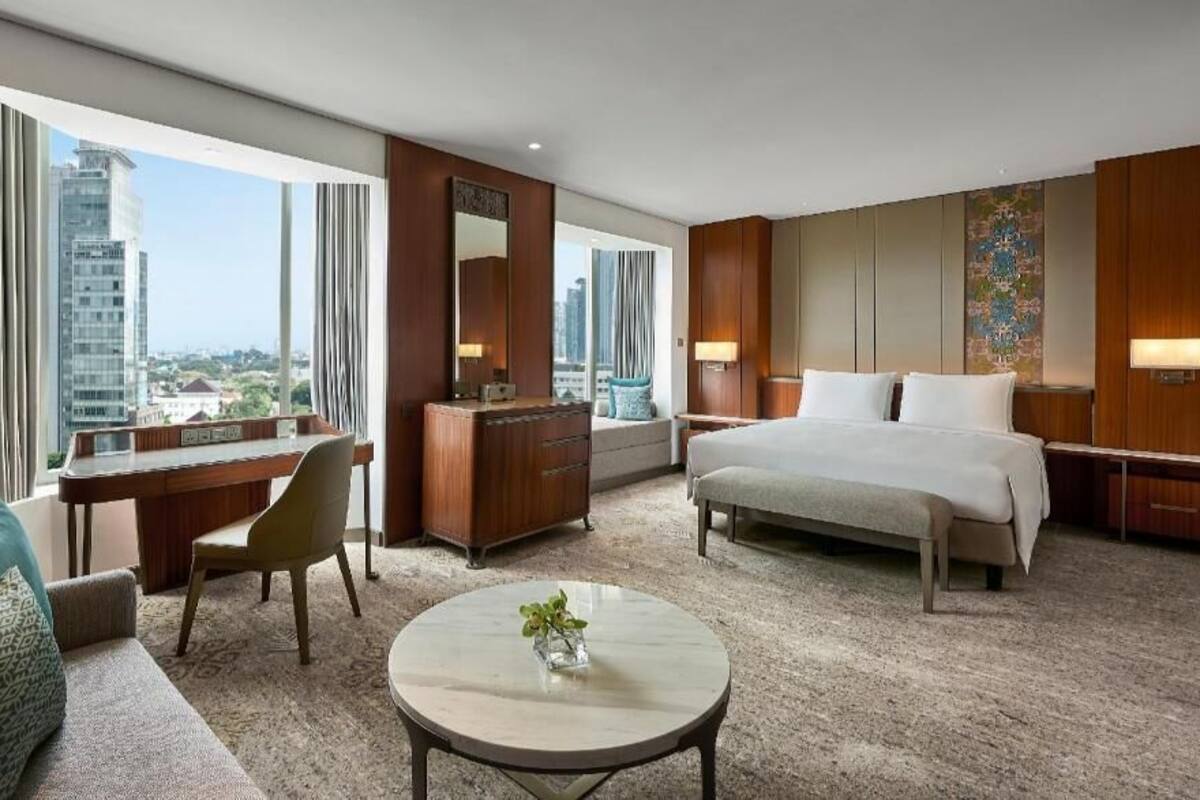 A hotel room with a large bed, a desk, a chair, a round coffee table, and a view of the cityscape.