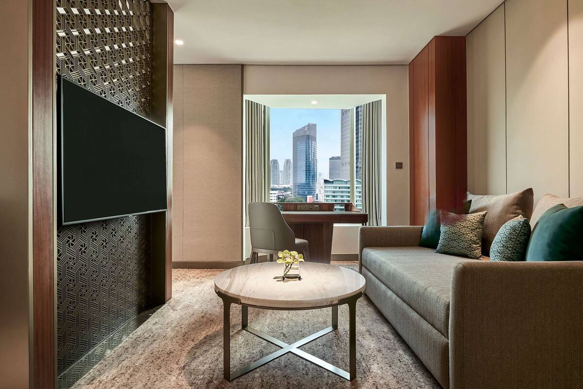 A modern hotel room with a city view, a flat-screen TV, a round coffee table, a sofa, and a desk with a chair.