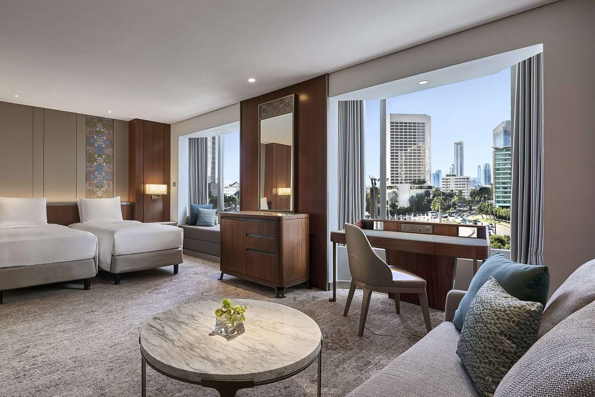 A hotel room with two beds, a desk, a chair, a coffee table, and a large window with a city view.