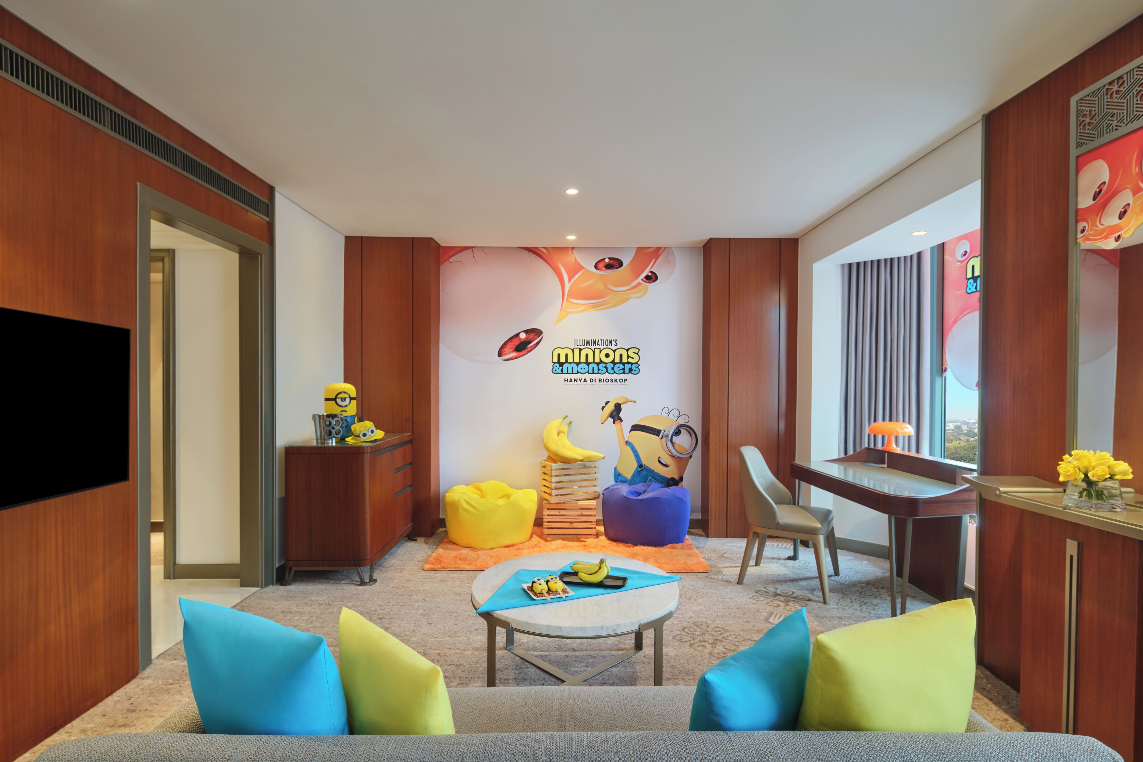 Habitación de hotel con un mural temático de Los Minions, un televisor de pantalla plana, un escritorio con lámpara y una zona de estar con cojines coloridos.