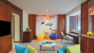Habitación de hotel con un mural temático de Los Minions, un televisor de pantalla plana, un escritorio con lámpara y una zona de estar con cojines coloridos.