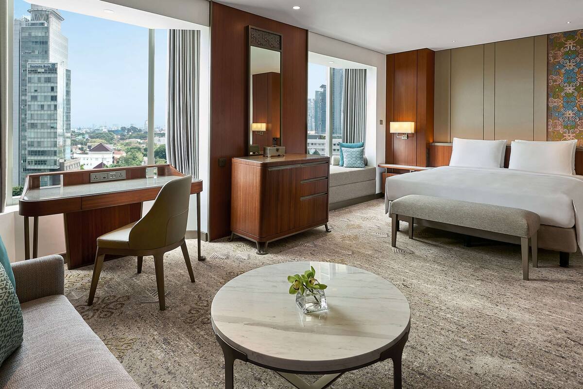 A hotel room with a large bed, a desk with a chair, a small table with a plant, and a view of the cityscape.