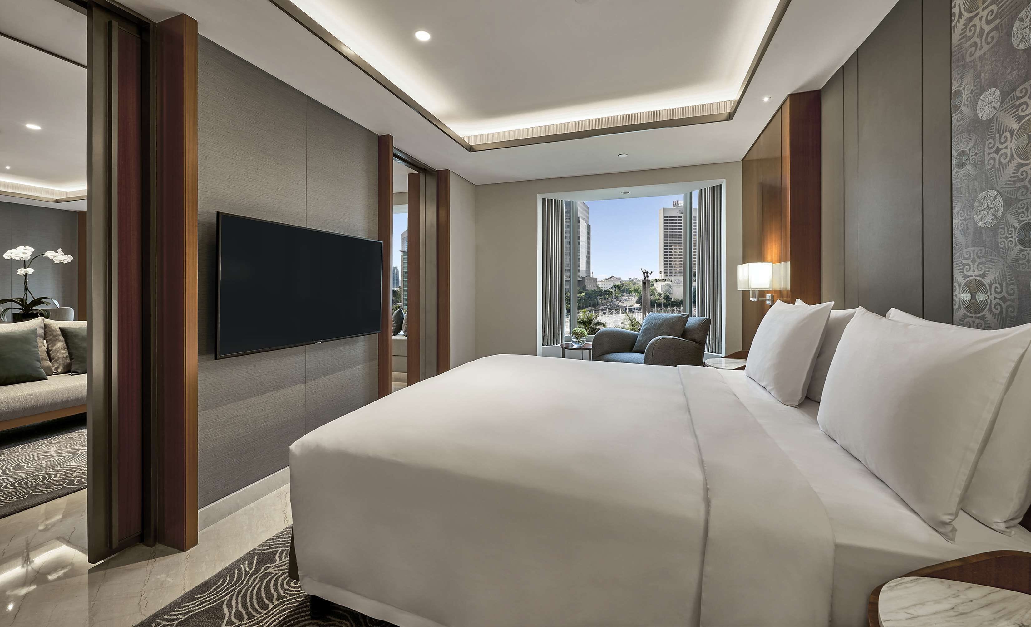 Quarto de hotel moderno com uma cama grande, televisão de tela plana na parede, área de estar com sofá e poltronas, e vista para a cidade através de janelas amplas.