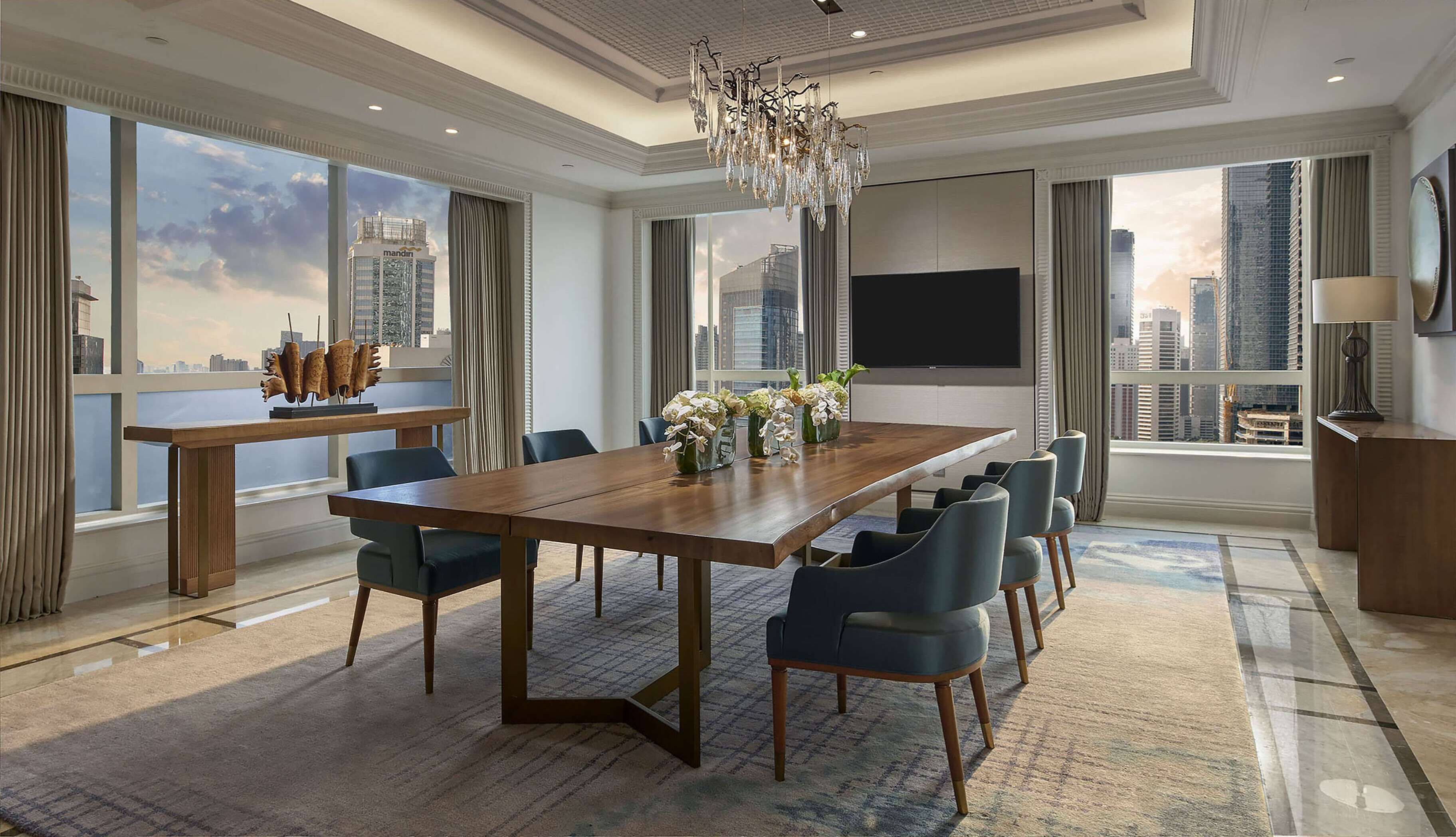 Una habitación de hotel moderna con una amplia mesa de comedor, asientos cómodos, un candelabro, un televisor de pantalla plana y vistas a la ciudad.