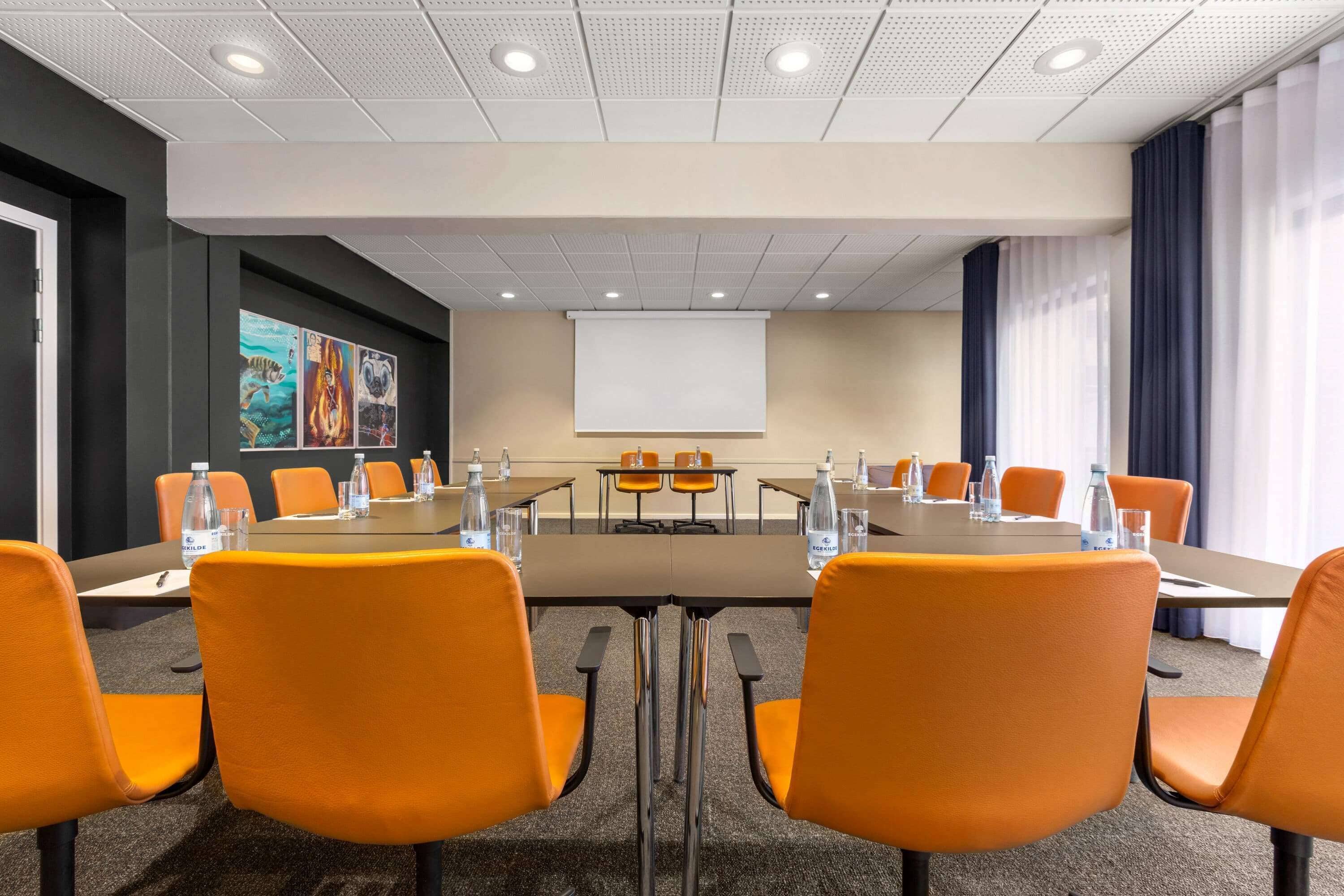 A conference room with orange chairs, a large table, a projector screen, and a water dispenser.