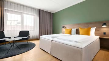 A hotel room with a bed, a desk, two chairs, a window with curtains, and a green wall.