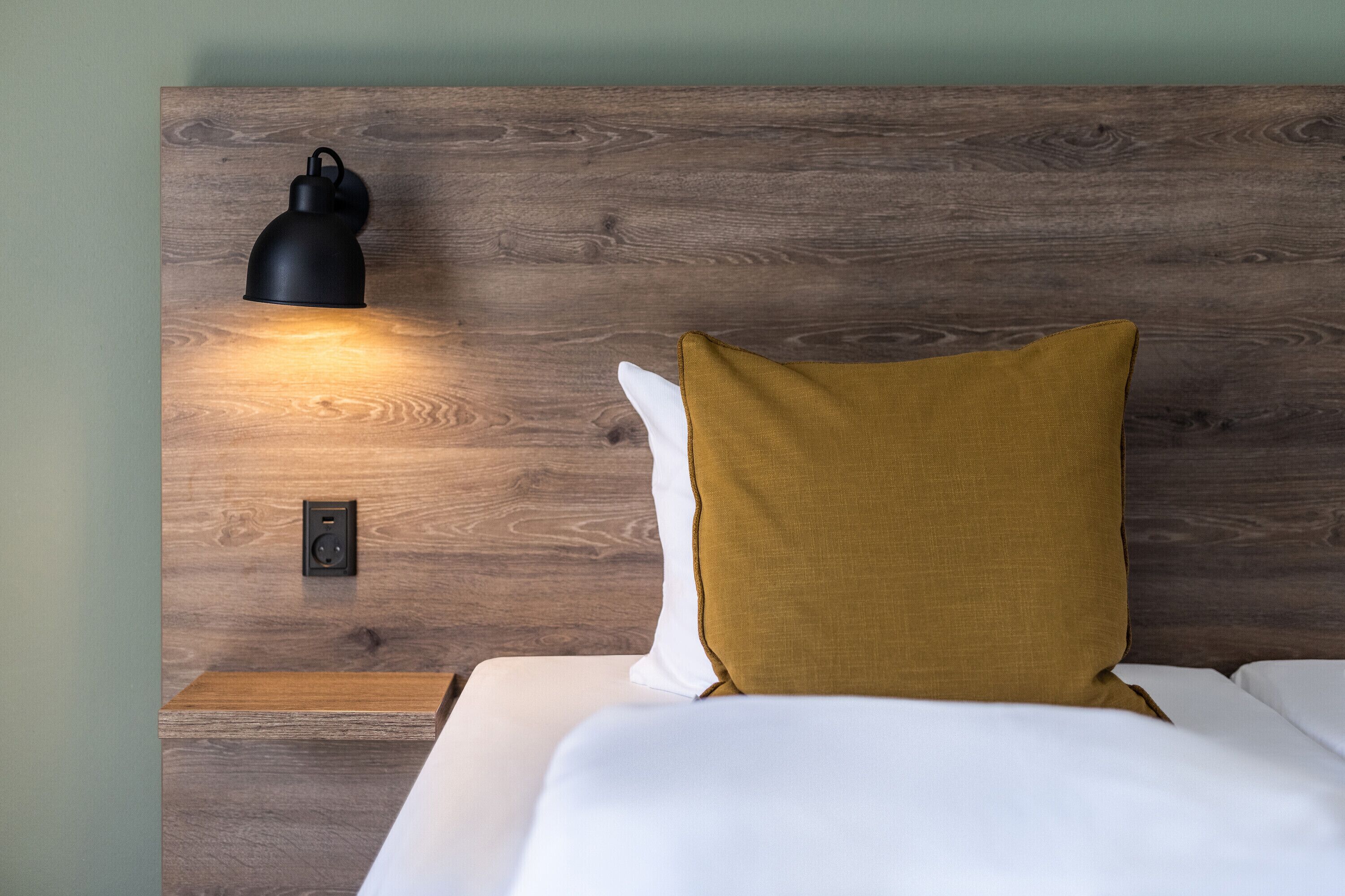 A bed with white bedding and a mustard-colored pillow against a wooden headboard with a wall-mounted lamp.