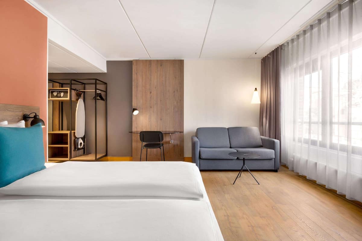 A hotel room with a bed, a grey sofa, a desk with a chair, and a wardrobe.
