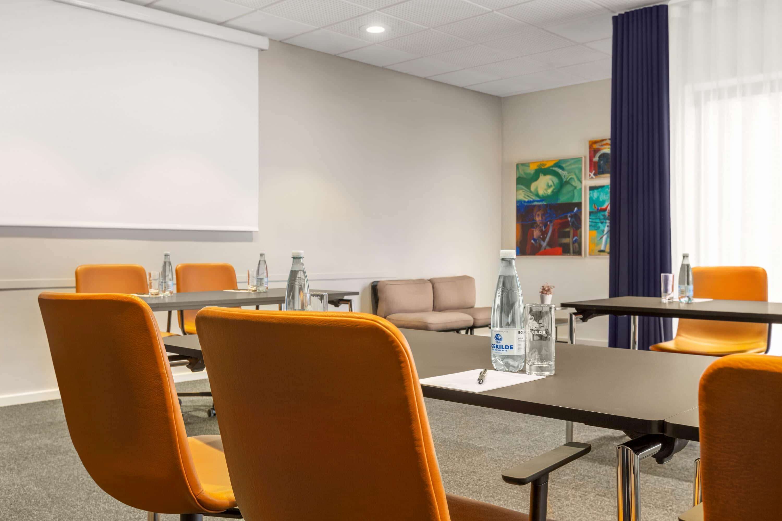 A conference room with a large table, orange chairs, a projector screen, and artwork on the wall.