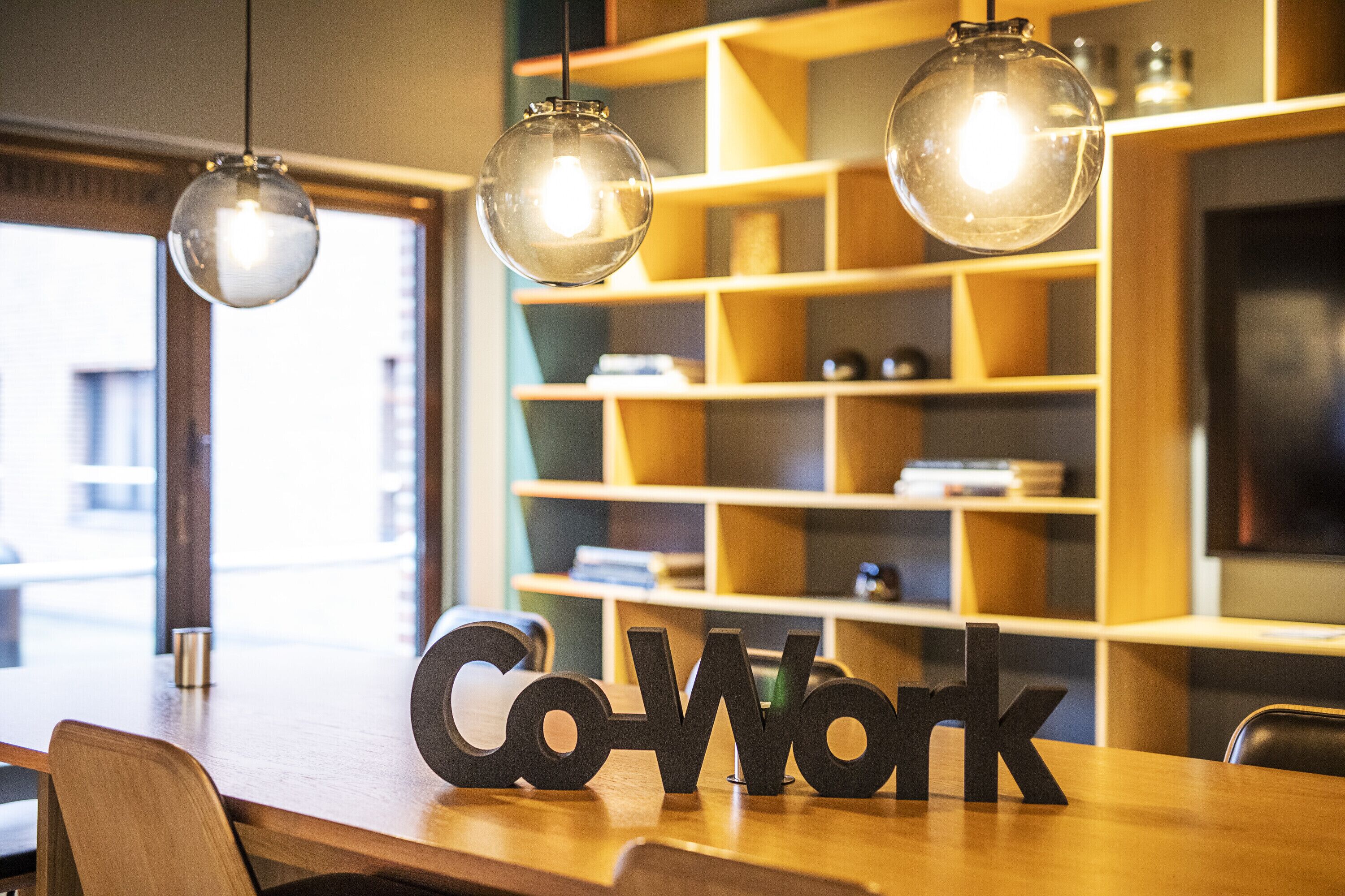 A wooden table with the words "Co-Work" in black letters, a bookshelf in the background, and two hanging pendant lights.